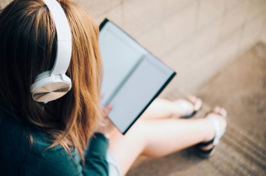 Gözlüklü gülümseyen kadın kitap okuyor ve kulaklıkla müzik dinliyor. Dışarıda, güneş ışığına karşı, şehir yaşam tarzı konseptine karşı..