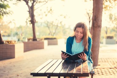 Gözlüklü gülümseyen kadın kitap okuyor ve kulaklıkla müzik dinliyor. Dışarıda, güneş ışığına karşı, şehir yaşam tarzı konseptine karşı..