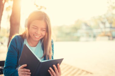 Gözlüklü gülümseyen kadın kitap okuyor ve kulaklıkla müzik dinliyor. Dışarıda, güneş ışığına karşı, şehir yaşam tarzı konseptine karşı..