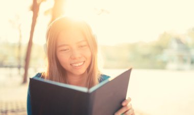 Gözlüklü gülümseyen kadın kitap okuyor ve kulaklıkla müzik dinliyor. Dışarıda, güneş ışığına karşı, şehir yaşam tarzı konseptine karşı..