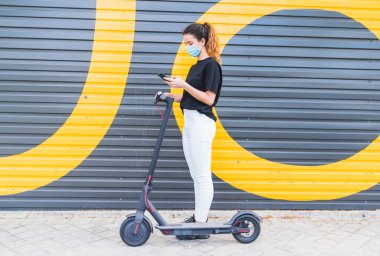 Yeni normal. Cep telefonu uygulamasını kullanarak elektrikli scooter 'ı çalıştıran maskeli bir kız. Elektrikli scooter kullanan maskeli bir genç. Genç bayan elektrik ve ekolojik taşıma kullanıyor.