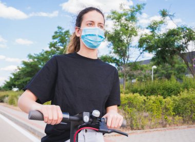 Yeni normal. Elektrikli scooter kullanan maskeli genç bir kız. Kadın elektrik ve ekolojik taşımacılık güvenliği ve salgın konseptini kullanıyor. Coronavirus cihazları. Sosyal mesafe. Coronavirüs