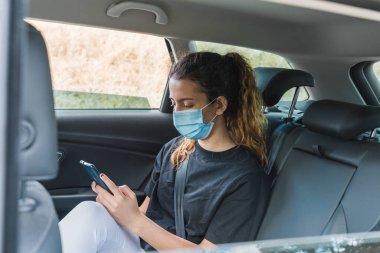 Yeni normal. Maskeli bir arabanın arkasında oturan bir ergen. Taksideki genç bir kadın maskeyle korunuyor. Güvenlik ve salgın konsepti. Coronavirus cihazları. Sosyal mesafe. Akıllı telefon kullanan genç kız.