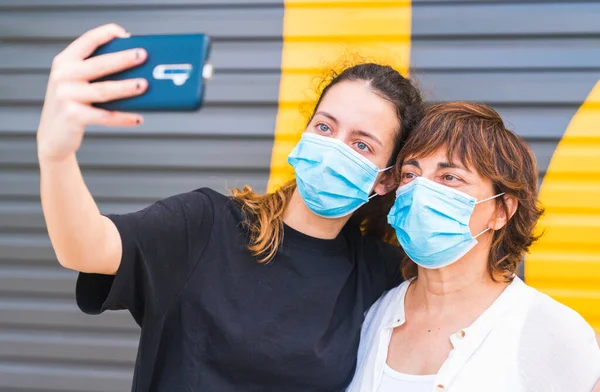 Anne ve kızı sokakta bir maske ile korunan selfie çekiyorlar. Aile maskeyle korunuyor. Maskeli aile özçekimi. Maskeli selfie. Sosyal mesafede insan hayatı. Yeni normal.