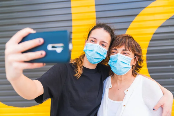 Anne ve kızı sokakta bir maske ile korunan selfie çekiyorlar. Aile maskeyle korunuyor. Maskeli aile özçekimi. Maskeli selfie. Sosyal mesafede insan hayatı. Yeni normal.