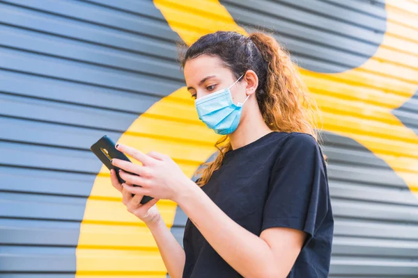Maskeli sokakta akıllı telefonunu kullanan bir ergen. Maskeyle korunan genç bir kadın. Sosyal mesafede insan hayatı. Güvenlik ve salgın konsepti. Coronavirus. Sosyal mesafe. Yeni normal.