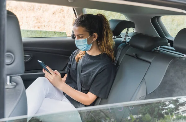 Yeni normal. Maskeli bir arabanın arkasında oturan bir ergen. Taksideki genç bir kadın maskeyle korunuyor. Güvenlik ve salgın konsepti. Coronavirus cihazları. Sosyal mesafe. Akıllı telefon kullanan genç kız.
