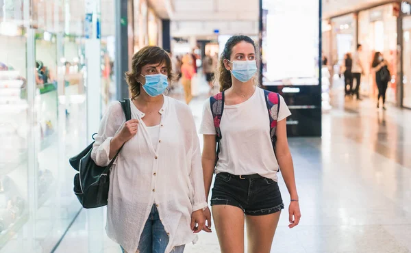 Maskeli bir anne ve kızı alışveriş merkezinden birlikte geçiyorlar. Maskeli kadınlar. Yeni normal. Yeni normalin maskeli eğlencesi. Alışveriş merkezinde maskeyle alışveriş yapmak.