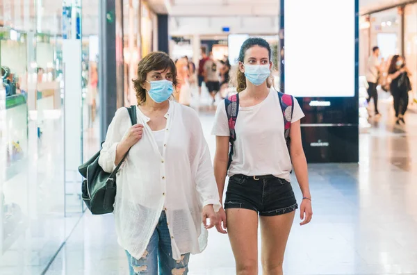 Maskeli bir anne ve kızı alışveriş merkezinden birlikte geçiyorlar. Maskeli kadınlar. Yeni normal. Yeni normalin maskeli eğlencesi. Alışveriş merkezinde maskeyle alışveriş yapmak.