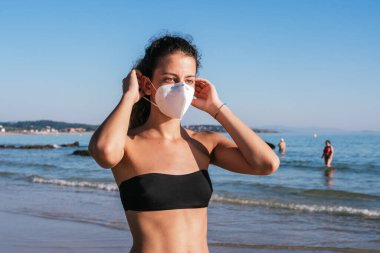 Maskeli, bikinili genç bir kız. Sahilde koronavirüs maskesi takıyordu. Halka açık yerlerde tıbbi maske takmak. Coronavirus karantinası. Covid-19 salgınında seyahat ve tatil.
