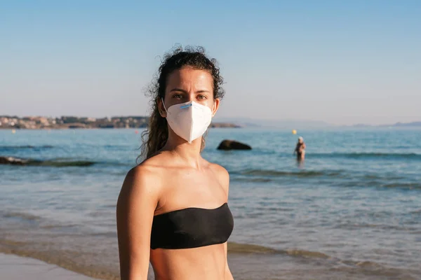 Young girl in a bikini with a mask. Wearing the coronavirus mask on the beach. Wearing the medical face mask in public places. Coronavirus quarantine. Vacation and travel in the Covid-19 pandemic.