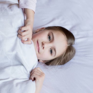 Yatakta tatlı küçük bir kız sabah uyandı. Çocuk yatağa uzanıyor ve yorganın altından dışarı bakıyor. Karantina sırasında evde vakit geçirmek. Ev konforu.