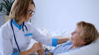 Genç bir kadın doktor, hastane yatağında yatan yaşlı bir hastanın elini tutuyor. Hastalık ve iyileşme sürecinde rahatlık ve destek sağlıyor.
