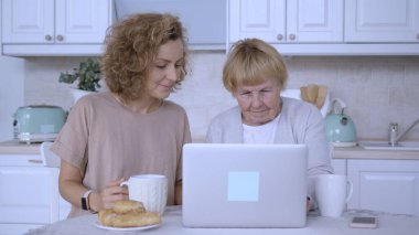 Yaşlı kadın mutfağında dizüstü bilgisayar kullanırken kızı onu izliyor ve çay içiyor. İkisi de gülümsüyor ve birlikte vakit geçiriyorlar.
