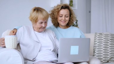 Gülümseyen bakıcı, elinde sıcak bir bardak tutan neşeli bir son sınıf öğrencisiyle dizüstü bilgisayarında bilgi paylaşıyor. İkisi de evdeki kanepede sıcak bir anın tadını çıkarıyor.