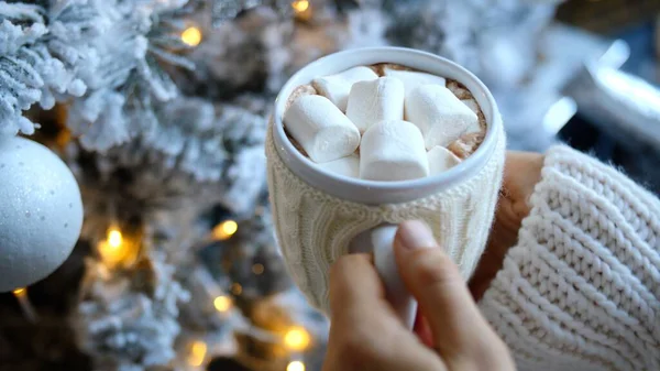 Beyaz yün kazak giyen kadın elinde bir kupa sıcak çikolata ve yanında süslemeli noel ağacı ve beyaz süslemeli şekerlemeler tutuyor.