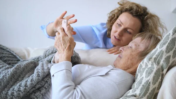 Hastane yatağında yatarken akıllı telefon kullanan yaşlı bir kadına yardım eden hemşire, hasta bakımını ve modern sağlık hizmetlerine teknolojik entegrasyonu gösteriyor