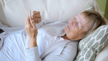 Kanepede rahat bir şekilde uzanan, rahat bir ifadeyle akıllı telefon kullanan, huzurlu bir ev ortamında modern teknolojinin tadını çıkaran yaşlı bir kadın.