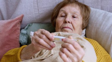 Soğuk algınlığı ya da soğuk algınlığı geçiren yaşlı bir kadın battaniyeyle kaplıyken termometreyi tutuyor, kendini kötü hissediyor, mevsimsel virüs, salgın hastalık...