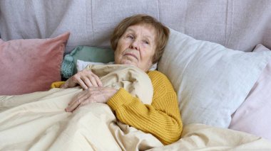Yaşlı kadın yatakta, battaniyelerle kaplı, sarı bir kazak giyiyor, düşüncelere dalmış, muhtemelen hayatını, deneyimlerini ve yaşlanma sürecini yansıtıyor.