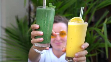 Güneş gözlüklü ve beyaz tişörtlü genç bir kadın elinde yeşil ve sarı renkli iki smoothie tutuyordu. Arka planda metal pipetler ve tropik yapraklar vardı.