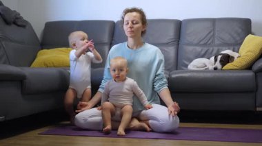 Genç anne mor minderde ikiz bebeklerle yoga yapıyor. Yakınlarda dinlenen küçük bir köpek. Huzurlu bir aile refahı ve samimi bir ev bağlantısı gösteriyor.