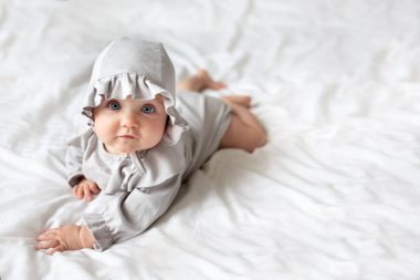 Evdeki yatağın üzerinde açık renk tulumlu küçük bir kız bebek kameraya bakıyor. Battaniyeye uzanmış sağlıklı bir çocuğun portresi. Metin için boşluk.