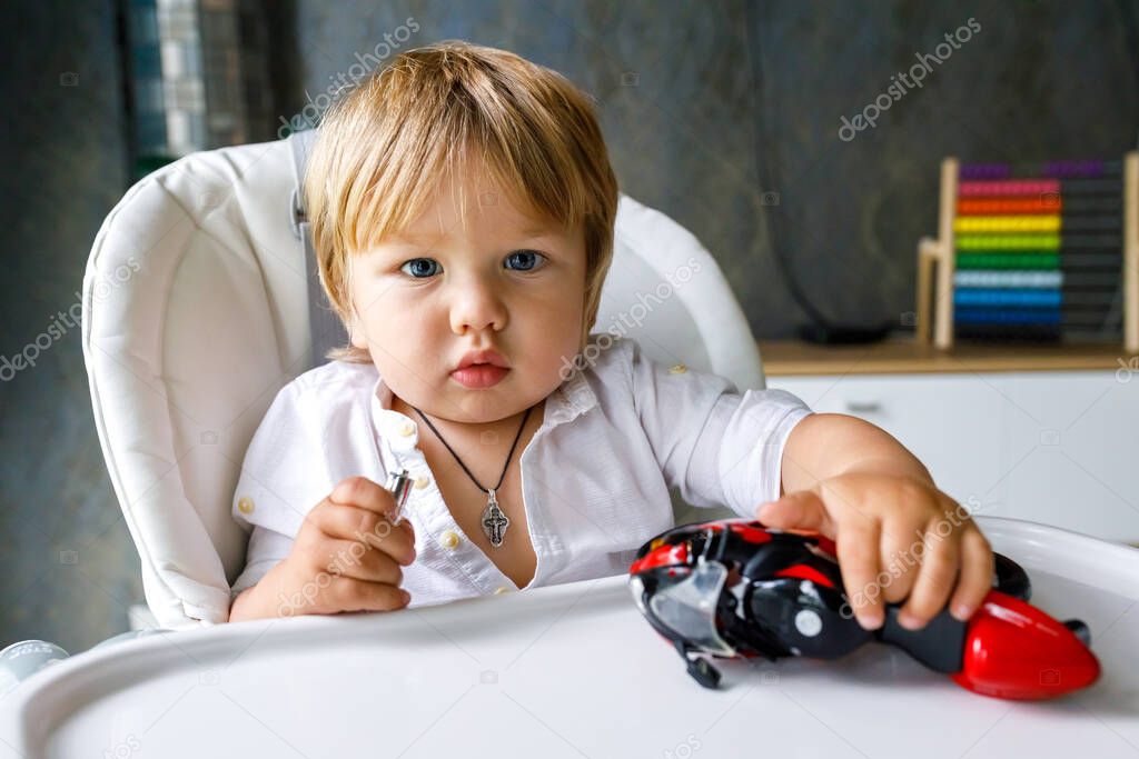 A cute little boy of European appearance is sitting on a white chair and playing with a toy motorcycle. The concept of child development at home