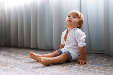 Evde güzel, sağlıklı bir çocuk yerde oturuyor ve şaşkınlıkla yukarı bakıyor. Evde yeni yürümeye başlayan sarışın bir çocuk. Metin için alanı kopyala