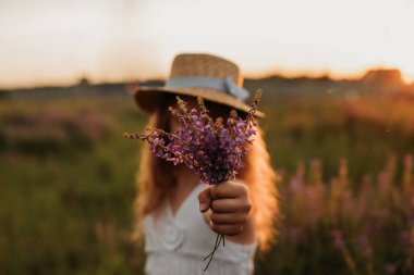 Hasır şapkalı bir kız yazın gün batımında bir tarlada duruyor ve bir demet mor çiçek gösteriyor.