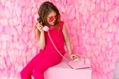 Küçük bir kız pembe gözlükleriyle tüylerinin pembe arka planında oturuyor. Güzel bir kız bebek eski pembe telefona şaşkınlıkla bakıyor. Metin için boşluk