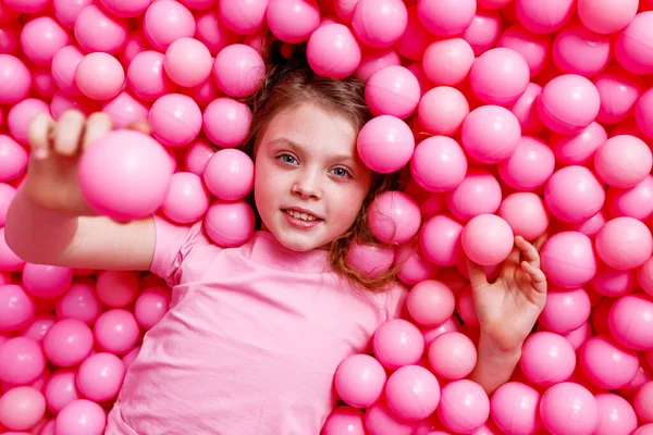 A little happy girl in pink clothes with a smile lies in pink plastic balloons on a pink background. The view from the top.