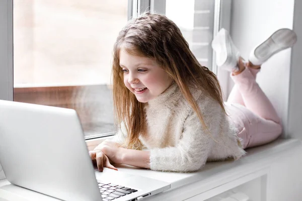 A beautiful girl with a laptop at home at the window looks at the screen and smiles. The girl at home communicates on the Internet. Kids distance learning.