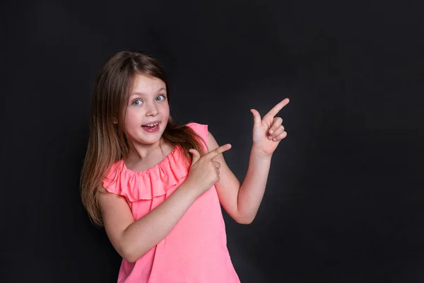 A happy surprised girl points to the right with her hands with her fingers up. Happy child on a black background. Copy space for text