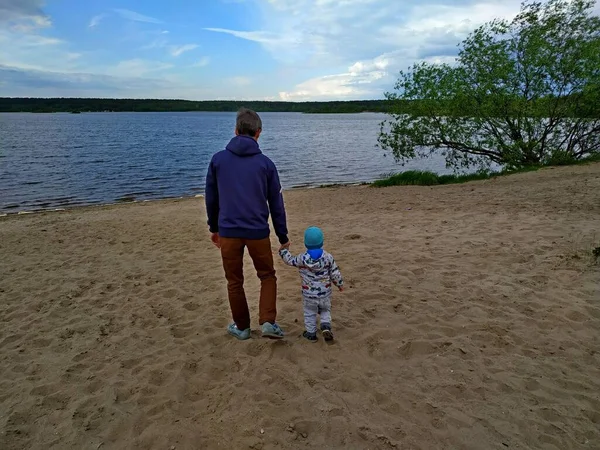 dad in a blue sweatshirt and brown trousers walks by the hand with his two-year-old son in the sand on the shore of a large lake. father and son. caring family. Fathers day. children protection day