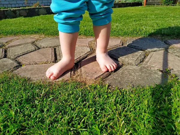 A small child in blue pants and barefoot stands on tile on a green lawn — Stock Photo, Image