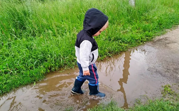 A kisfiú gumicsizmában sétál át a pocsolyán. gondtalan nyár. Jogdíjmentes Stock Képek