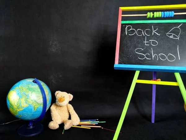 Невелика дошка, кольорові олівці, глобус і плюшевий ведмідь на чорному. Назад до школи — стокове фото