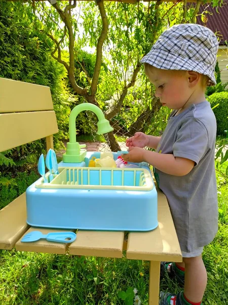 Niño aprende a lavar los platos en un lavabo de juguete en el patio trasero —  Fotos de Stock