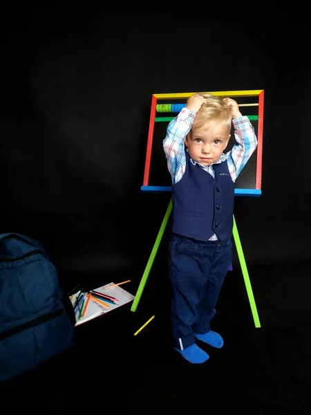 Niño pequeño con un traje azul sostiene su cabeza mientras está de pie junto a la pizarra —  Fotos de Stock