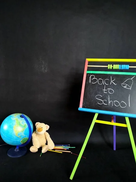 Невелика дошка, кольорові олівці, глобус і плюшевий ведмідь на чорному. Назад до школи — стокове фото