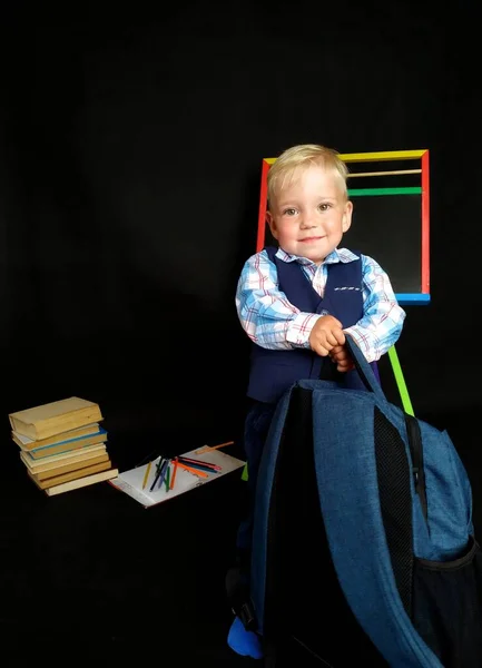 Niño sonriente en un traje azul sostiene gran mochila mientras está de pie junto a la pizarra —  Fotos de Stock