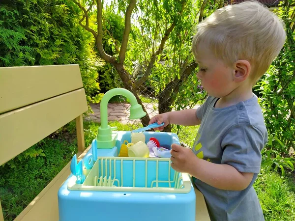 Niño aprende a lavar los platos en un lavabo de juguete en el patio trasero —  Fotos de Stock