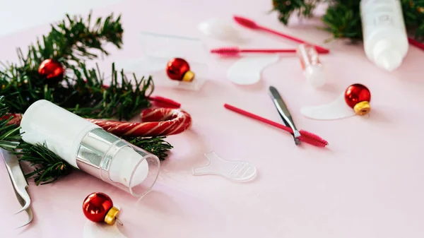 Güzellik salonu ve kirpik sanatçısı aletleri ve tedavi ürünleri sol tarafta, Noel temalı pembe arka planda bir çerçeve oluşturuyorlar. Şeker ve Noel oyuncakları ve tedavi araçları