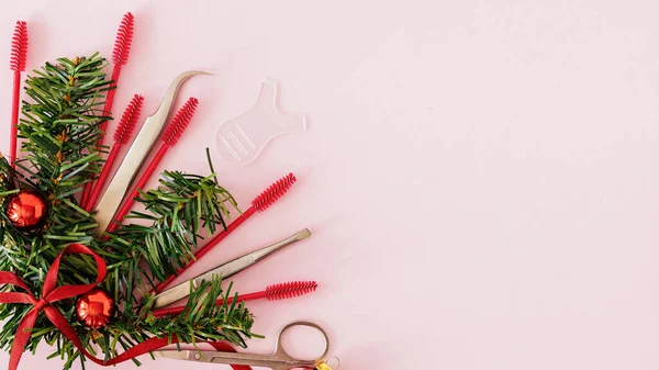 Güzellik salonu ve kirpik sanatçısı aletleri ve tedavi ürünleri sol tarafta, Noel temalı pembe arka planda bir çerçeve oluşturuyorlar. Tedavi için aletleri olan Noel ağacı oyuncakları. Boşluğu kopyala.