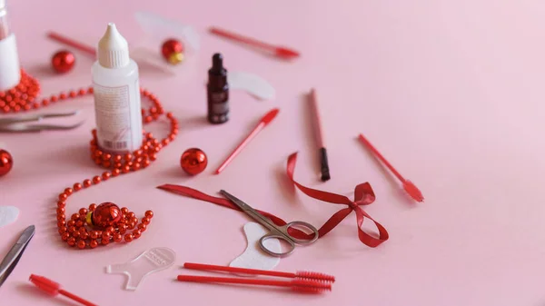 Güzellik salonu ve kirpik sanatçısı aletleri ve tedavi ürünleri sol tarafta, Noel temalı pembe arka planda bir çerçeve oluşturuyorlar. Tedavi için aletleri olan Noel ağacı oyuncakları. Boşluğu kopyala.