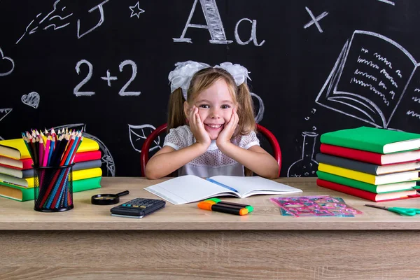 Colegiala sonriente sentada en el escritorio con libros, útiles escolares, sosteniendo sus dos manos sobre las mejillas —  Fotos de Stock