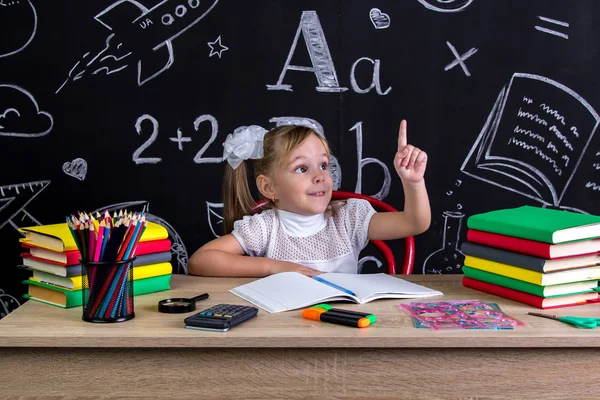 Colegiala sentada en el escritorio con libros, útiles escolares, sosteniendo su dedo hacia arriba. Se me ocurre el pensamiento correcto. — Foto de Stock