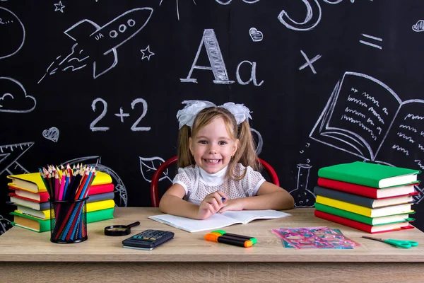 Colegiala emocionada sentada en el escritorio con libros, útiles escolares, sosteniendo la pluma en la mano derecha — Foto de Stock