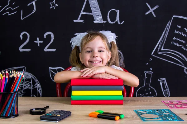 Una colegiala sonriente sentada en el escritorio con un montón de libros debajo de la barbilla, rodeada de útiles escolares. Pizarra como fondo —  Fotos de Stock